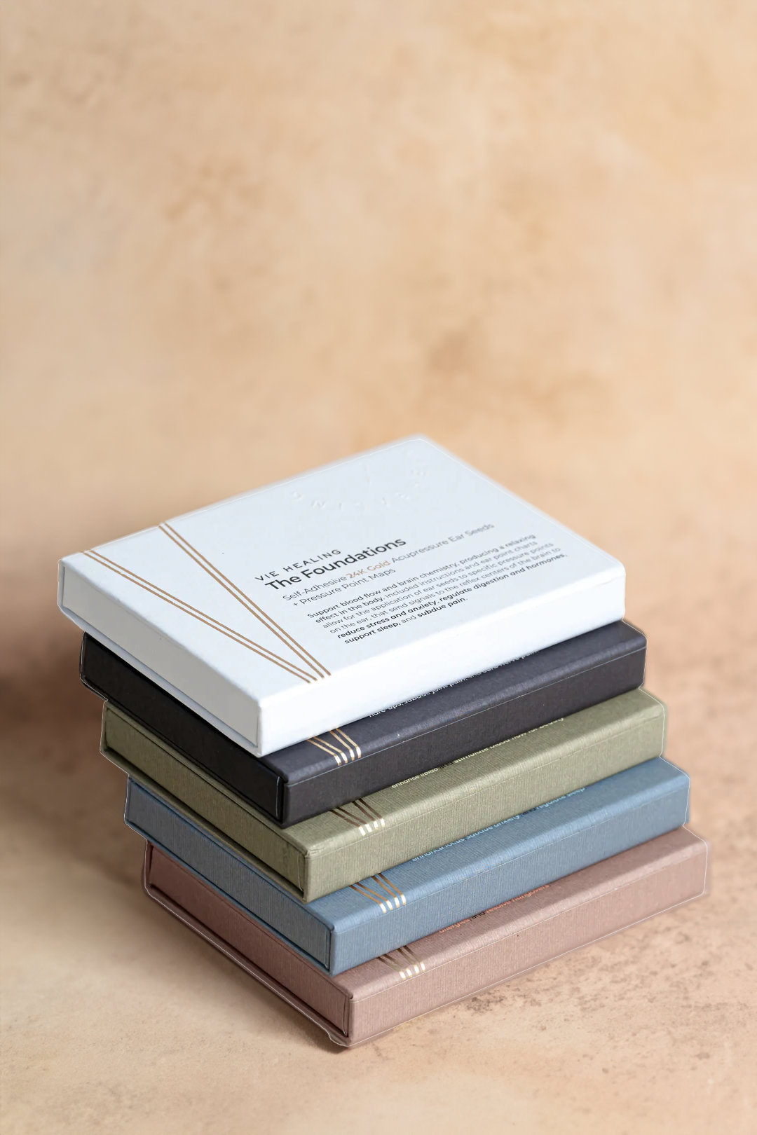 Stack of VIE HEALING 24K gold ear seed sets, including The Foundations, in earthy linen-textured boxes