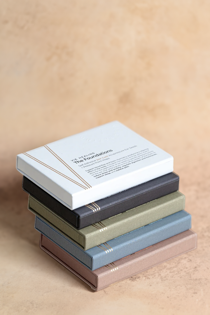 Stack of VIE HEALING 24K gold ear seed sets, including The Foundations, in earthy linen-textured boxes