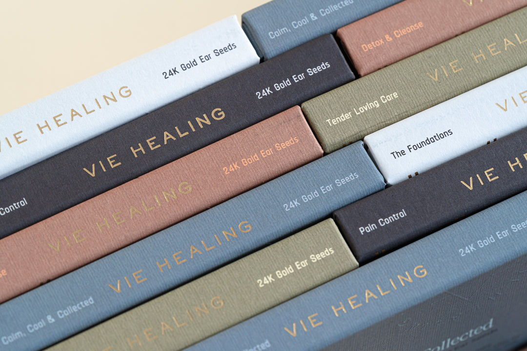 Close-up of VIE HEALING 24K Gold Ear Seeds boxes showing themed sets — Calm Cool & Collected, Detox & Cleanse, Tender Loving Care and Pain Control